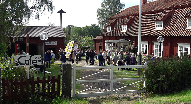 Stort rött hus med människor utanför på gårdsplanen. En skylt med texten Café.