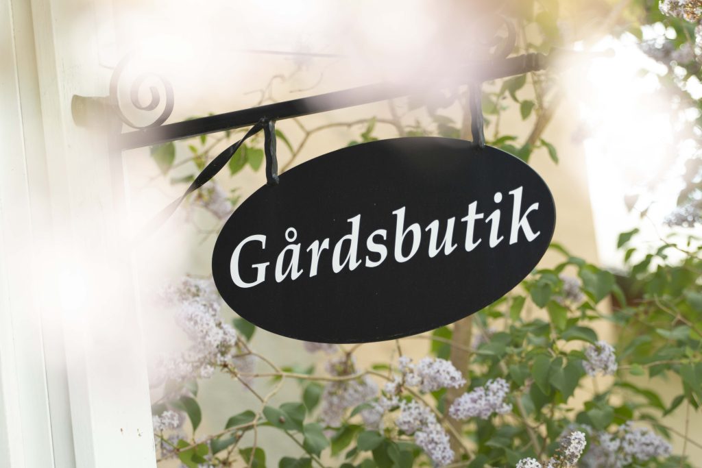 Skylt med texten Gårdsbutik framför blommande syrenbuske.